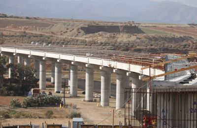 The motorway under construction is pictured with visible rebar and concrete structures, highlighting the project's ongoing progress.